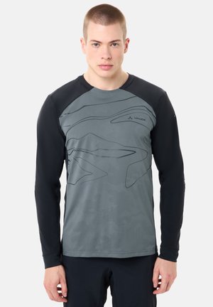 Jonge man met kort haar, gekleed in een grijs-zwart sportshirt met lange mouwen en abstracte lijnen, gecombineerd met een donkere broek, staand tegen een witte achtergrond.
