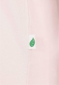 Jasnoróżowy materiał z pionową szwem i małą białą metką z zielonym ikoną liścia naszytą na szwie.