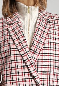 Blazer a quadri rosa e bianco con dettagli neri, caratterizzato da rever affilati. Sotto, è visibile un maglione zip-up a coste crema.