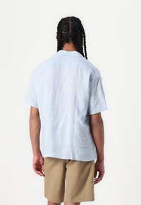 Himmelblaues kurzärmliges Hemd mit vertikalen weißen Streifen, lockerer Passform und einem Yoke-Detail auf dem Rücken. Getragen mit beigen Shorts.