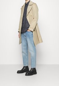 Cappotto beige con design doppiopetto, jeans dritti blu chiaro e stivaletti neri in pelle con suola spessa. Camicia a trama sotto.