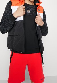 Hombre vistiendo un chaleco acolchado color bloque negro y naranja, sudadera negra con estampado y pantalones cortos rojos brillantes con bolsillos con cremallera negra.