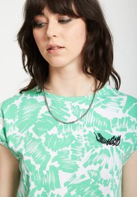 Groen en witte abstracte penseelstreken t-shirt met een relaxed fit, ronde halslijn en zwarte geborduurde accent op de borst. Zilveren kettinghalsketting zichtbaar.