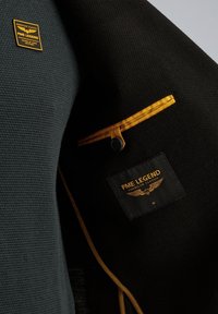 Zwarte textuurjack met een gladde binnenvoering, voorzien van een gele accent, een logo patch en een knoopsluiting. Maatlabel zichtbaar.