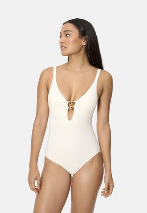 Mujer con un traje de baño blanco de una pieza con tirantes finos y dos adornos de anillos dorados en el pecho, mirando hacia un lado sobre un fondo liso.