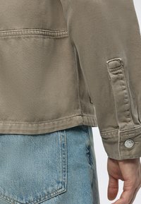 Gros plan d'une personne portant une manche de veste beige et un jean en denim bleu clair avec des coutures visibles et un bouton sur le bord de la manche de la veste.