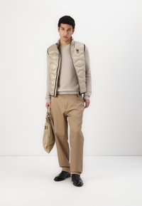 Gilet trapuntato beige sopra un maglione leggero in maglia, abbinato a pantaloni a gamba larga color cammello e scarpe nere. Tiene una borsa beige, con elementi di design minimalisti.