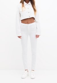 Chaqueta de mezclilla blanca recortada con dos bolsillos en el pecho y botones plateados, combinada con jeans blancos ajustados y zapatillas blancas.