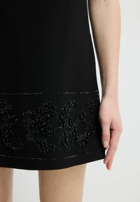 Czarna sukienka z teksturowaną kwiatową haftem ozdobionym koralikami i cekinami wzdłuż brzegu. Gładka tkanina i dopasowana sylwetka.