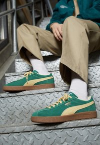 Baskets en daim vert avec des accents beiges et des semelles en caoutchouc, dotées de lacets et d'un logo, posées sur un escalier en tôle métallique diamantée.