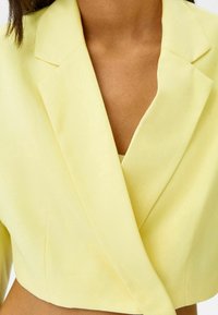 Zkrácený blazer v jemně žluté tkanině, s klopovým límcem, čistými liniemi a mírným leskem. Minimalistický design s dobře padnoucím střihem.