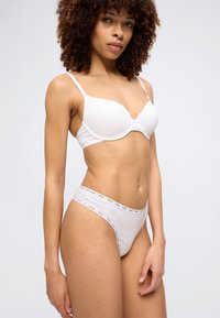 Femme aux cheveux bouclés portant un soutien-gorge blanc et une culotte blanche taille haute assortie avec une ceinture élastique de marque, debout devant un fond uni.