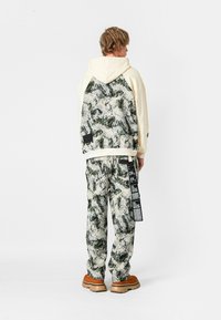 Sudadera con capucha en crema con un respaldo estampado en verde y beige; pantalones a juego con el mismo estampado y etiquetas laterales. Unos zapatos marrones completan el conjunto.