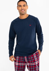 Marineblauses Hemd mit Raglanärmeln und einem kleinen roten Emblem. Kombiniert mit rot-blauen karierten Pyjamahosen. Aus weichem Baumwollmaterial.