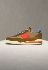 Καφέ και καμηλό αθλητικό παπούτσι με κόκκινη πλαϊνή επιφάνεια, μπλε ταμπόν στη φτέρνα, λευκά και μπλε κορδόνια, και λευκή σόλα με πορτοκαλί διαβαθμισμένη λεπτομέρεια.