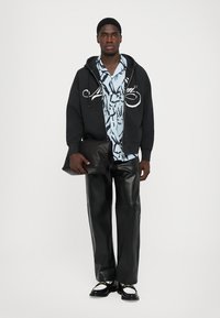Felpa nera con cappuccio e grafica bianca, camicia azzurra a fantasia sotto, pantaloni in pelle neri e pochette nera. Scarpe nere con accenti bianchi.