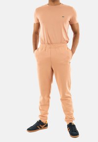 Camiseta de manga corta color melocotón con un pequeño logo verde, combinada con pantalones jogger a juego que cuentan con una cinturilla elástica. Zapatillas negras con acentos naranjas.