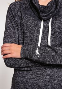 Huvtröja med cowl-krage i mörkgrått stickat tyg med vita snörningar, med en figurnära siluett och en logotypdetalj på ärmen.