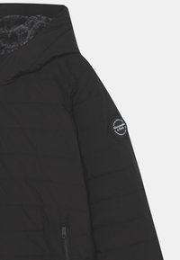 Schwarze Steppjacke mit Kapuze, ausgestattet mit einem weichen grauen Innenfutter, horizontalen Nähten und einem runden schwarzen Logopatch am Ärmel.