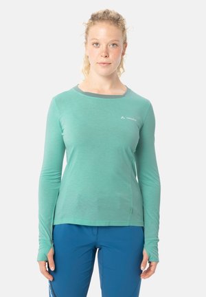 Donna con capelli biondi ricci che indossa una maglia a maniche lunghe color verde acqua e pantaloni blu, in piedi di fronte a uno sfondo semplice.