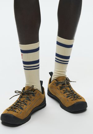 Dunkelhäutige Beine, die cremefarbene Socken mit marineblauen Streifen und hellbraune Wanderschuhe mit schwarzen Sohlen und blau gemusterten Schnürsenkeln auf weißem Hintergrund tragen.