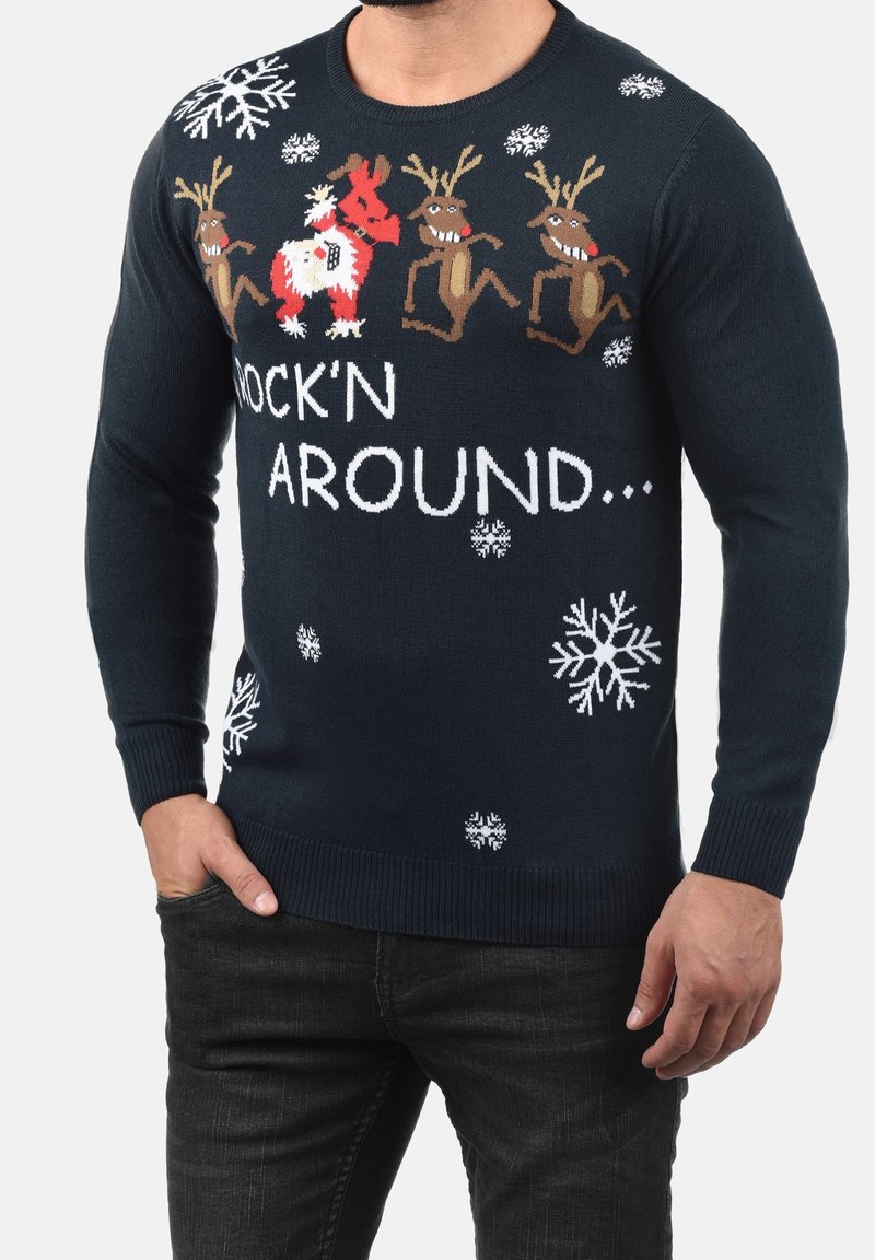 Pull de Noël bleu marine avec des rennes dansants, le Père Noël, des flocons de neige blancs et le texte « Rock'n Around », porté par un homme les mains dans les poches.