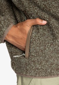 Brun, strukturerad fleecepuls med en framficka. Fodret är slätt och beige, med en dragsko-detalj vid nederkanten.