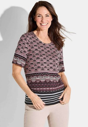Glimlachende vrouw met lang bruin haar die een kortemouwen patroon top met strepen en cirkels draagt, staand tegen een witte achtergrond.