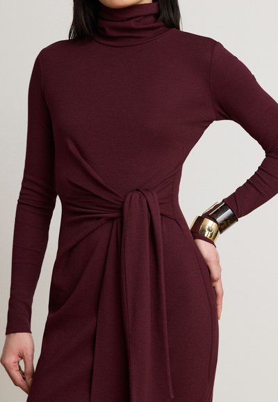 Femme portant une robe bordeaux à manches longues avec un col montant et un détail noué à la taille, accessoirisée d'un large bracelet manchette métallique.