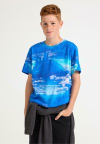 Kortærmet T-shirt med et grafisk design af blå himmel og skyer. Blødt stof, afslappet pasform. Kombineret med mørke jeans, grå sweater knyttet om taljen.