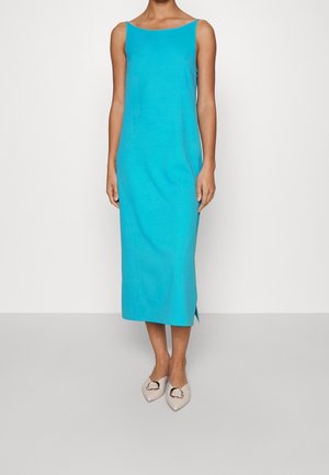 Femme portant une robe midi turquoise sans manches avec une fente latérale, assortie à des ballerines beiges à bout pointu ornées de détails en boucle dorée.