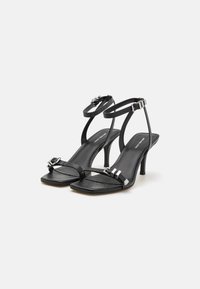 Sandales à talons en cuir noir avec de fines brides autour de la cheville et à l'avant du pied, bout carré, accents métalliques et un talon aiguille modeste.
