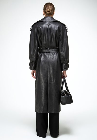 Persoon met haar in een knot, gekleed in een zwarte leren trenchcoat met ceintuur en zwarte broek, die een klein zwart handtasje naast zich houdt.