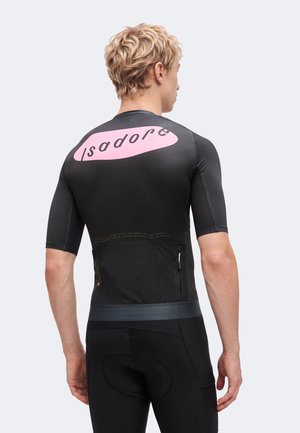 Blonde man draagt een zwart wielertenue met een roze "Isadore" logo op de bovenrug, met de rug naar de kijker en handen ontspannen langs het lichaam.