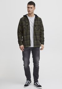 Grønternet flannel skjorte med sort hætte, to brystlommer, knaplukning foran. Båret over en grå t-shirt og parret med mørke jeans og sorte sneakers.