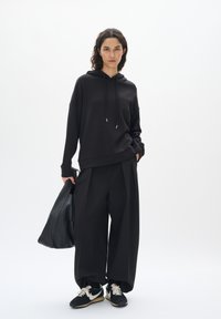 Felpa nera con cappuccio e lacci, abbinata a pantaloni neri larghi. Il modello tiene una borsa nera. Le sneakers presentano una combinazione di nero e bianco.