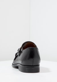 Chaussure habillée en cuir noir à texture lisse, bout rond et une seule lanière à boucle. Présente un talon bas et des détails cousus.