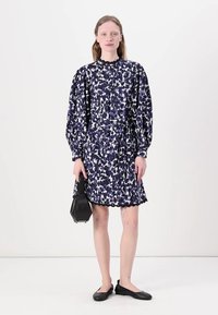 Blumenmusterkleid in Marineblau und Weiß mit Puffärmeln, tailliertem Schnitt und gewelltem Saum. Kombiniert mit einer kleinen schwarzen Handtasche und Ballerinas.