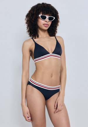 Femme aux cheveux bouclés portant des lunettes de soleil blanches à monture œil de chat et un bikini bleu marine avec une bordure rayée rouge, blanche et noire, debout devant un fond clair.