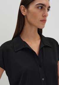 Zwarte T-shirt met korte mouwen en een kraag, voorzien van een knopenpat met een gladde, matte textuur. Eenvoudig, strak ontwerp zonder patronen.