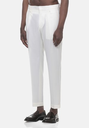 Uomo che indossa pantaloni bianchi su misura con risvolti e mocassini neri in pelle, in piedi davanti a uno sfondo chiaro e uniforme.