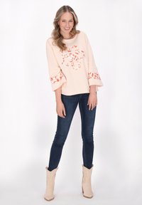 Helles rosa Oberteil mit floralem Design, Glockenärmeln, kombiniert mit dunkelblauen Jeans und beigen Cowboystiefeln. Glatte Textur, lässiger Stil.