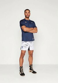 Αθλητικός άντρας στέκεται με τα χέρια σταυρωμένα, φορώντας μπλε UFC μπλούζα, λευκές σορτς με σχέδια και μαύρα ψηλά μποτάκια πυγμαχίας.