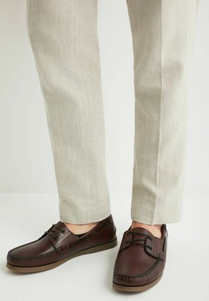 Brązowe skórzane buty typu boat shoes noszone z jasnobeżowymi spodniami, widoczne od połowy łydki w dół na jednolitym białym tle.