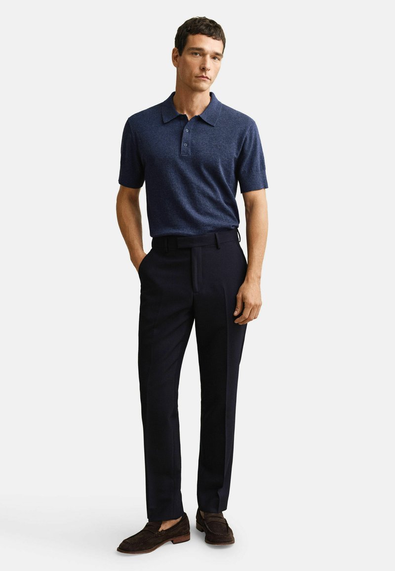 Homme debout, main dans la poche, portant un polo à manches courtes bleu marine, un pantalon noir et des mocassins marron foncé, sur fond blanc.