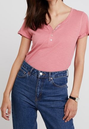Kvinde iført en kortærmet lyserød henley-skjorte indstukket i højtaljede blå jeans, med et sort armbåndsur på venstre håndled.