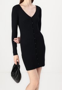Robe en tricot côtelé noir avec un décolleté en V, des manches longues et une fermeture à boutons sur toute la longueur. Associée à un sac en cuir noir.