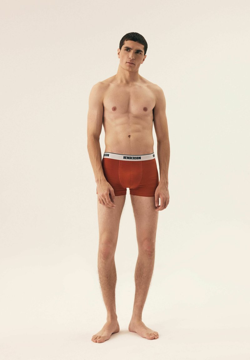 Czerwone bawełniane bokserki z białym elastycznym pasem, na którym widnieje napis "HENDERSON". Model stoi boso, pozując w pozycji wyprostowanej na neutralnym tle.