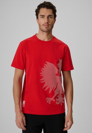 Mężczyzna z krótkimi kręconymi włosami w czerwonej koszulce z dużym białym wzorem graficznym na dolnej prawej stronie, stojący na jednolitym tle.