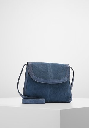 Lille marineblå ruskind crossbody taske med klaplukning og tynd skulderrem på en hvid piedestal mod en ensfarvet grå baggrund.
